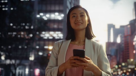 Portrait of a Beautiful Japanese Female Walking in Smart Casual Clothes and Using Smartphone in a City at Sunset. Stylish Woman Connecting with People Online, Messaging and Browsing Internet.