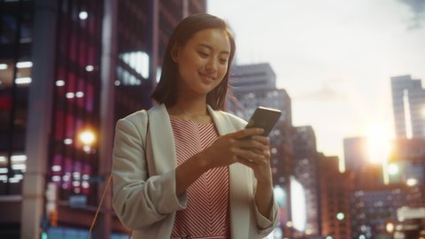 Portrait of a Beautiful Japanese Female Wearing Smart Casual Clothes and Using Smartphone in a City at Sunset. Stylish Woman Connecting with People Online, Messaging and Browsing Internet.