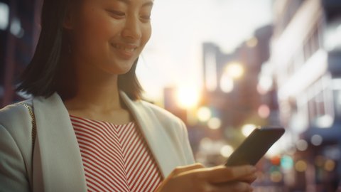 Close Up Portrait of an Attractive Japanese Female Wearing Smart Casual Clothes and Using Smartphone in a City at Sunset. Stylish Woman Connecting with People Online, Messaging and Browsing Internet.