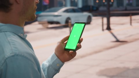 Close Up of a Young Man Using Smartphone with Green Screen Chroma Key Mock Up Display in Vertical Position on a Street During a Sunny Day. He's Tapping and Swiping the Screen.