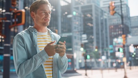 Portrait of a Handsome Young Man Walking in Casual Clothes and Using Smartphone on the Urban Street. Smiling Male in Big City Connecting with People Online, Messaging and Browsing Internet.