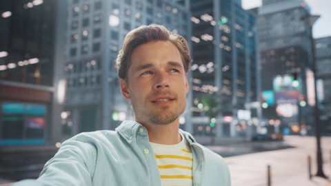 POV Video Call Portrait of a Handsome Young Stylish Man in Casual Clothes Talking to Friends or Colleagues while Standing Outside on City Street. Waving Hand in Urban Environment.