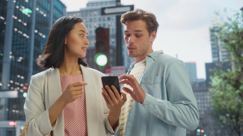 Young Stylish Multiethnic Couple Standing on a Street in a Big City. Attractive Japanese Female Showing Smartphone to Handsome Caucasian Male. Diverse Friends Enjoying Travelling Together.