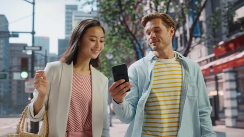 Young Stylish Multiethnic Couple is Casually Walking on a Street in a Big City. Handsome Caucasian Male Showing Smartphone to Beautiful Japanese Female. Diverse Friends Enjoying Travelling Together.