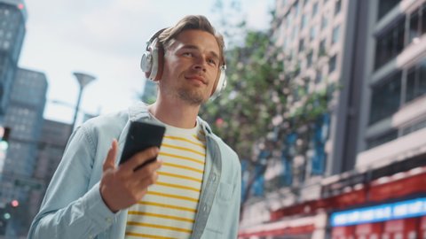 Portrait of a Handsome Young Man Casually Walking on the Street, Listening to Music on Headphones from Smartphone. He Uses Phone in Big City Messaging People Online and Browsing Internet.
