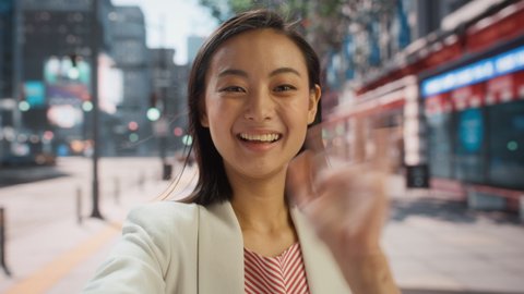 POV Video Call Portrait of a Beautiful Young Stylish Japanese Female in Smart Casual Clothes Talking to Friends or Colleagues while Standing Outside on City Street. Waving Hand in Urban Environment.