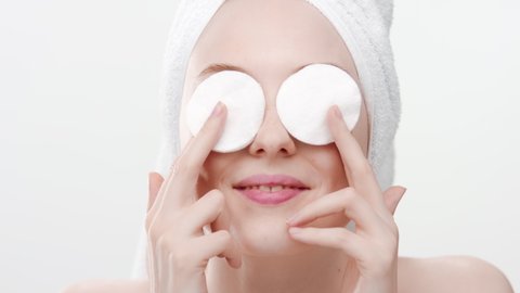 Funny gorgeous fit European young woman with hair towel on her head holds cotton pads on her eyes and smiles wide for the camera on white background | Eyelid care concept