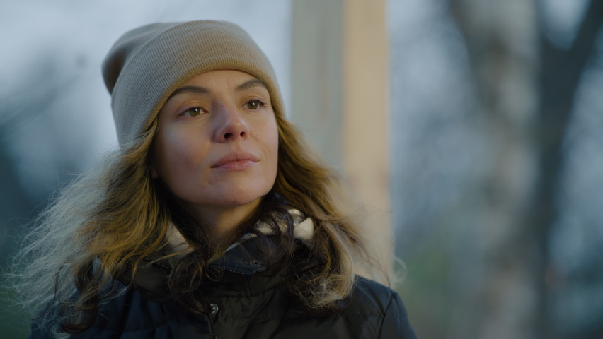 Close-up of a woman in a winter hat drinking hot tea from a mug on a cold autumn day, looking around pensively and sadly