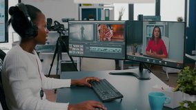 African woman editor with headset talking during online conference with partner on video call editing client work, getting feedback on commercial movie using post production software on pc in office