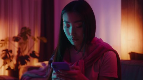 Beautiful asian young woman use phone sitting on sofa in neon light room at home. Joyful, homey, smartphone, chatting, attractive, youth, darkness, lady, alone, typing, tapping, resting. Slow motion