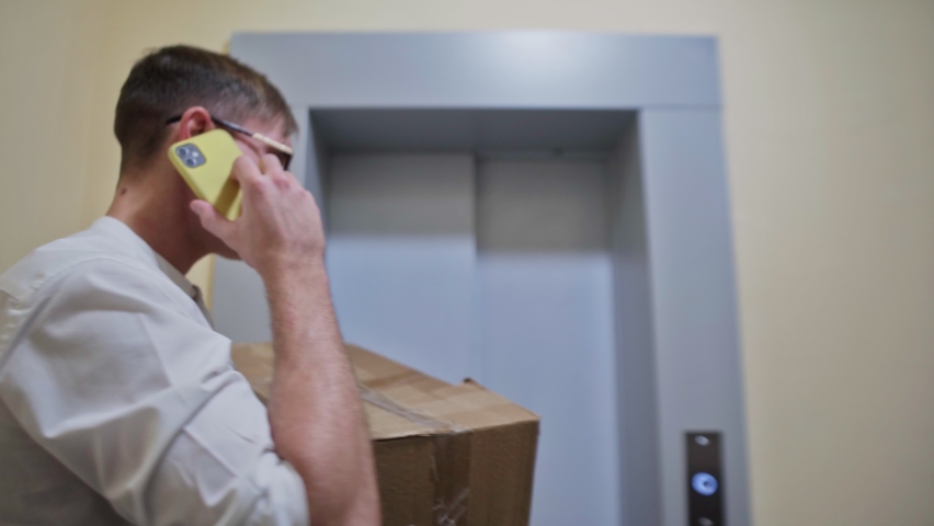 Young man courier in glasses uses phone, holds cardboard box and enters in the elevator. Man moving in to new appartment