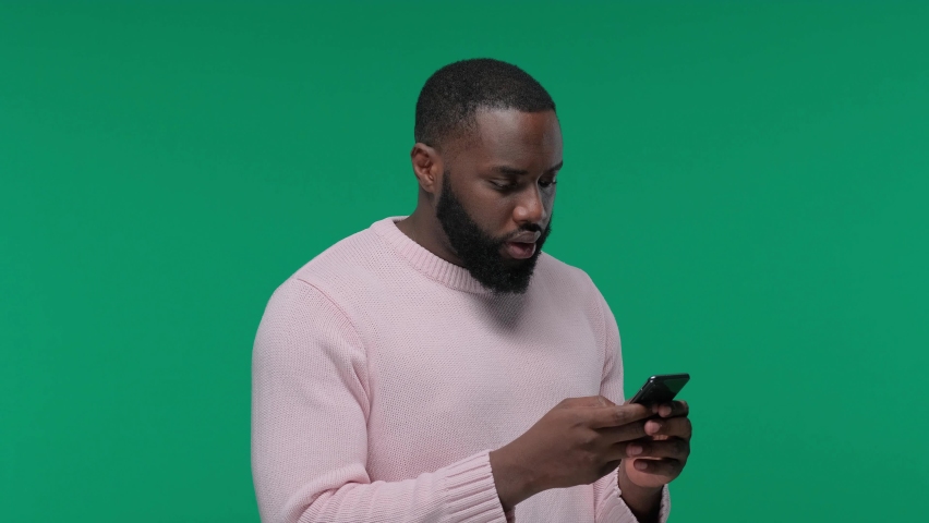 young black man excited and celebrating while holding and looking at his phone on Green Screen, Chroma Key Royalty-Free Stock Footage #1074857582