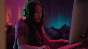 Asian esports girl playing online video game with friendly team. Beautiful concentrated woman in headphones speaker using keyboard and mouse. Home interior.