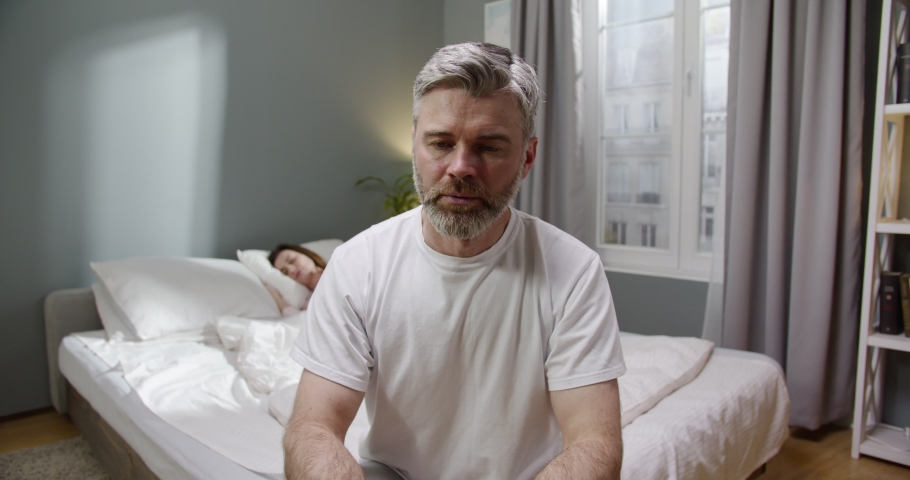 So upset. Waist up portrait view of the sad bearded man sitting at the bed and looking at his sleeping wife after the quarrel. Family disagreements concept Royalty-Free Stock Footage #1074863999