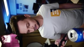 Young attractive bearded, brown-eyed, man vlogger with dark hair and mustache, dressed in grey t-shirt, is holding smartphone in hands and speaking, looking at camera. Vertical video frame 9x16