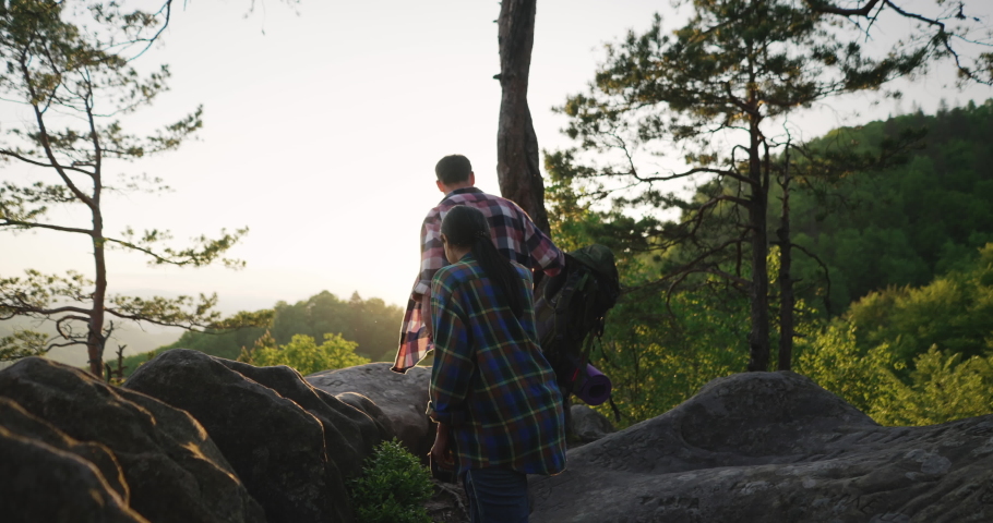 Romantic couple admiring sunset in mountains. Two travelers hikers reached the top of rocky hill, standing together, embracing and looking at bright evening sun light.
