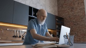 Cute 60-year-old gray-haired old man with beard at age of using computer browsing Internet, reading an e-book in application communicates online via video link with doctor. Distance learning old man.