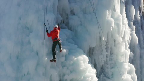 Стоковое видео: Aerial view of a frozen waterfall and rocks with climber ascending its ice-covered surface, using ice axes and crampons