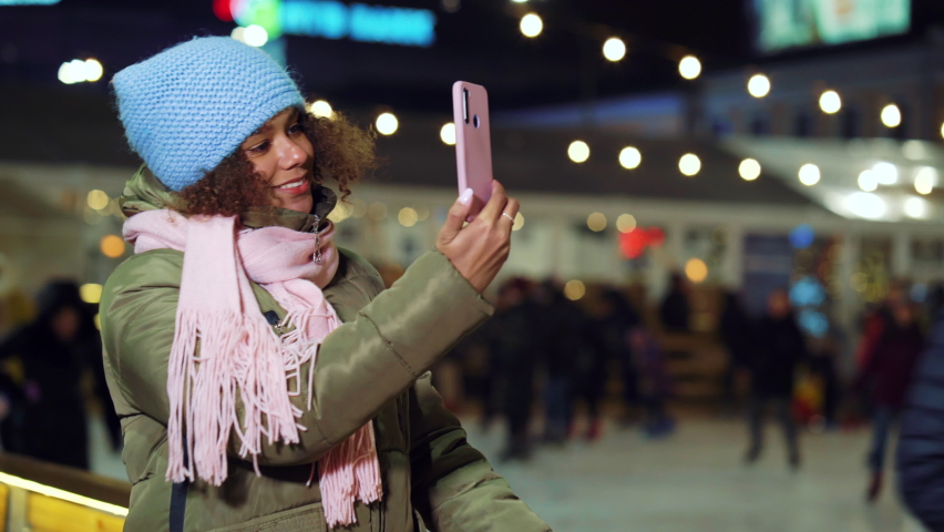 Black girl in winter outfit recording video of people skating on rink, blurred background. Young blogger streaming live from Christmas market. Concept of technology
