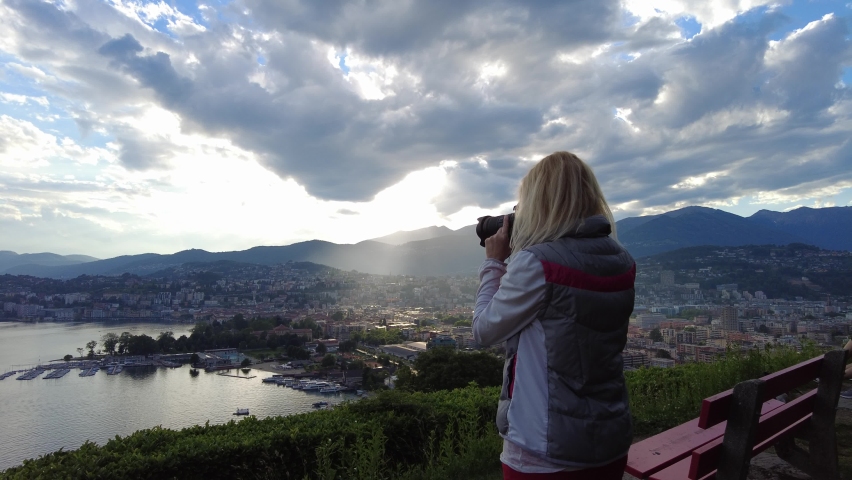 Woman photographer taking pictures on top of Lugano city by Lugano Lake in Switzerland. Lookout with an aerial view from San Michele hill. Lugano skyline with San Salvatore mount in Ticino canton.
