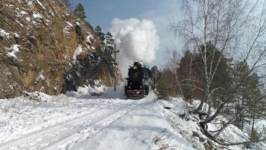 Aerial close to cinematic old authentic steam locomotive, engine rides on railroad railway and releases thick white clouds of steam from pipe. Winter snowy nature. Historical Soviet Union epic train