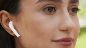 Closeup Of Brunette Woman Wearing Earbuds Listening To Music Looking Aside And Smiling Posing In Park Outdoors. Young Lady Enjoying Playlist Outside. Cropped, Side View