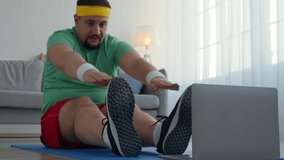Abdominal muscles stretching. Chubby man in bright clothes training at home, exercising against laptop on floor, practicing stretch exercise, close up, slow motion