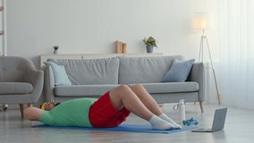 Hard sport training problem. Overweight man in bright sports clothes exercising at home, trying to do sit ups on floor, watching online video tutorial on laptop, slow motion