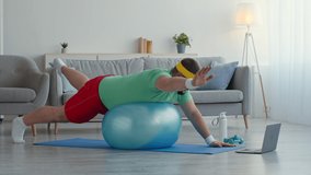Pilates for fat people. Overweight guy in bright clothes lying on fit ball and lifting his legs and hands, watching fitness lesson on laptop, doing stretching exercises at home, slow motion
