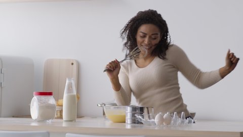 Cooking is fun. Young emotional african american woman singing with whisk and dancing, enjoying music, preparing food at kitchen alone, slow motion