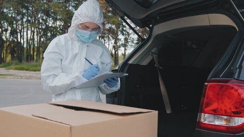 Female medical worker doctor courier in protective clothing white uniform latex gloves loads big cardboard boxes of medicines in trunk of car writes number of parcels on paper document counting goods