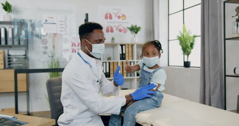 Handsome caring positive experienced black-skinned male doctor and cute small black-skinned girl both in protective masks posing on camera in modern medical office with thumbs up