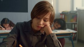 Tired schoolboy sitting in classroom. Cheerful boy boring during lesson at elementary school. Pensive kid holding hand on face. Smart student writing in notebook with classmates on background  - Powered by Shutterstock - Get 15% off with code: PIKWIZARD15