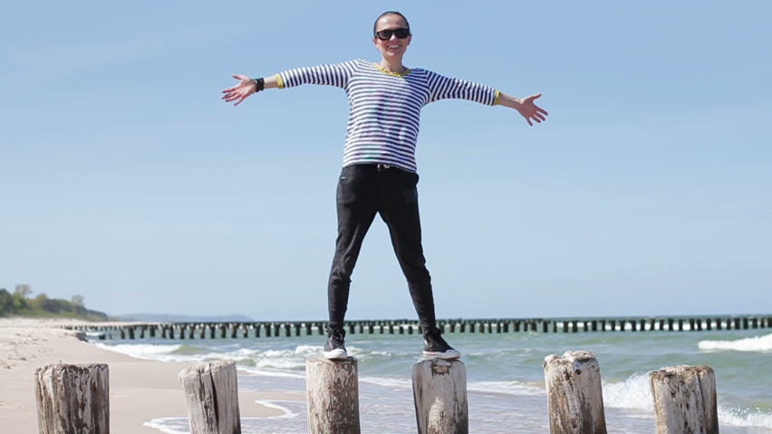 Woman standing on wooden breakwaters and raising arms to the sky on beach, feeling the breeze on sea background, Baltic Sea, slow motion