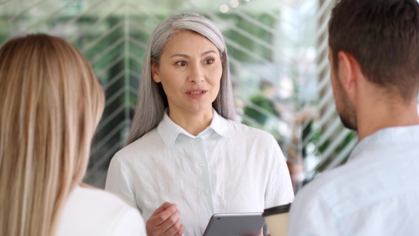 Asian senior businesswoman ceo sales agent mentor talking to young Caucasian male and female interns trainees discussing business project using tablet device standing in modern corporate office. Royalty-Free Stock Footage #1074905687