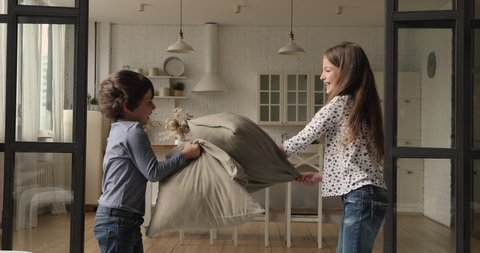 Crazy small child boy fighting pillows with laughing older sister in modern living room. Carefree little kids siblings best friends having fun entertaining playing cushion battle on weekend at home.