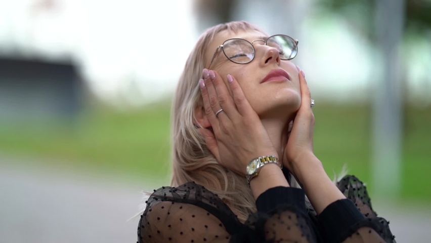 portrait of a blonde with glasses and a black blouse. she raises head, strokes neck, and hugs shoulders