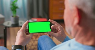Older man at Home Resting on a Couch Using with Green Mock-up Screen Smart Phone in Horizontal Landscape Portrait Mode. Woman Using Tablet Device, Browsing Internet, Watching Content, Videos.