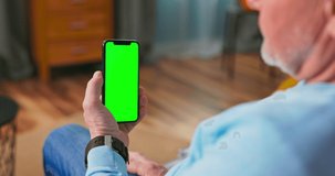 Senior Man at Home Uses Green Mock-up Screen Smartphone. She's Sitting On a Couch in His Cozy Living Room. Over the Shoulder Camera Shot