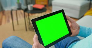 Close up of digital tablet with blank green mock up Screen display hold by old adult man relaxing in cozy room at home. Camera moves away from the tablet.