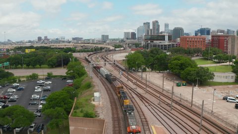 Aerial view of cargo train slowly riding through EBJ Union station. Backwards flying drone following railway corridor. Skyline with skyscrapers in background. Dallas, Texas, US