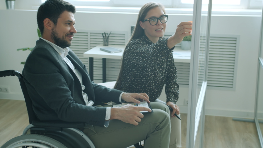 Colleagues disabled man and healthy woman discussing information on sticky notes working in office together looking at glass board and talking