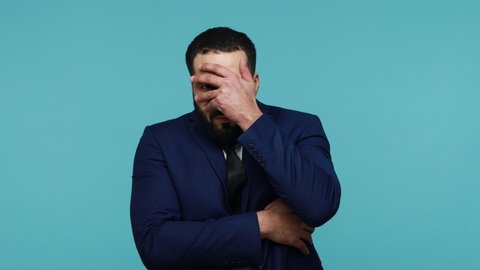 Brunette attractive curious young man with beard in official style suit closing eyes with palm, spying, taking glance looking through fingers. Indoor studio shot isolated on blue background.