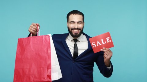 Joyful excited male with beard in business suit holds shopping bags and sale word, shouting from happiness, man satisfied with discounts and purchase. Indoor studio shot isolated on blue background.