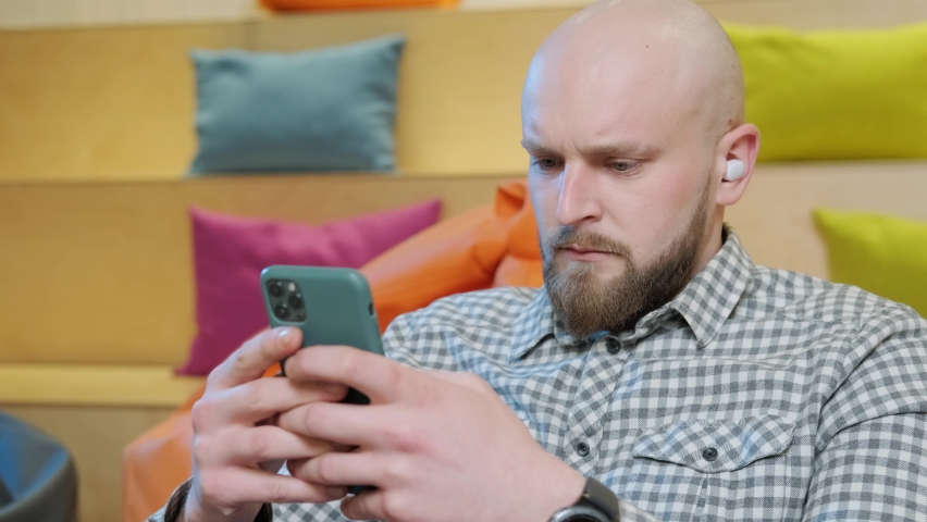 Close up of an young man sits on a bean bag while playing online using his phone. Spend time online during isolation at home, colorful background.