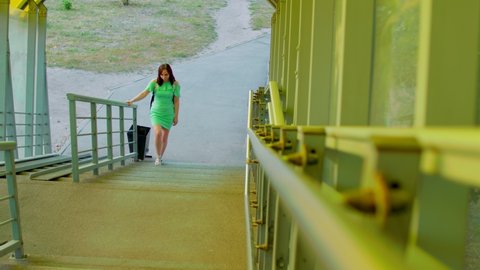 Front view of young woman in dress climbs pedestrian crossing. Brunette walking along overground pedestrian in summertime. Concept of safe transition above highway.
