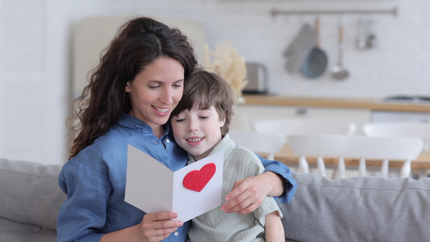 Caring son present mom greeting card for holiday event: mother day or birthday sit cuddling to mum reading postcard. Happy pleased young single mother hugging small boy on family holiday or valentines Royalty-Free Stock Footage #1074940724