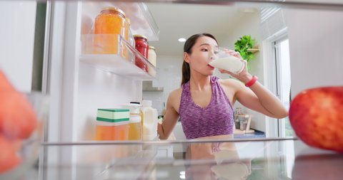 asian woman takes milk and drinks it from opened refrigerator in kitchen at home