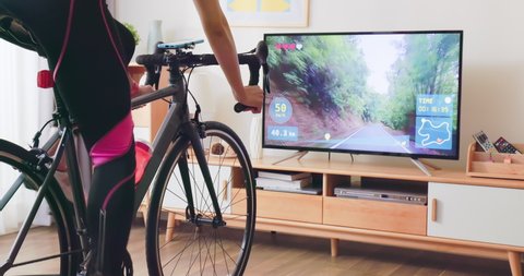 Asian woman cyclist is exercising in the house by cycling on trainer and play online bike games with TV