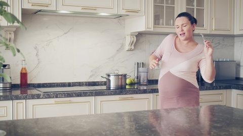 Mom listening music and relaxing stay at home. Cute fun mum dancing and preparing breakfast on kitchen. Concept pregnancy and happiness. Happy pregnant  woman singing in Kitchen Sunny Day.